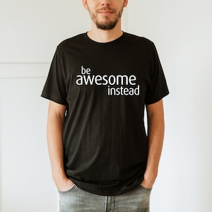May include: Black t-shirt with the white text "be awesome instead". The shirt is a crew neck style and is made of a soft material. The person is wearing gray jeans.