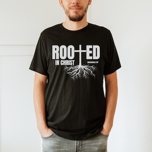 Puede incluir: Camiseta negra con la frase blanca "ROOTED IN CHRIST", una cruz y un diseño de raíces. El texto "Efesios 3:17" también está impreso en la camiseta. La camiseta es de cuello redondo.