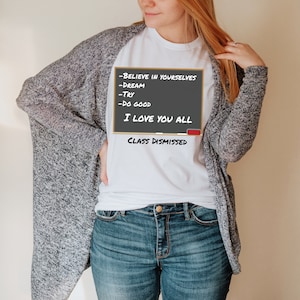 A woman wearing a white t-shirt that has a graphic of a blackboard on it. On the blackboard a lesson is written. It says, Believe in Yourselves, Dream, Try, Do Good. I Love You All. Class Dismissed.