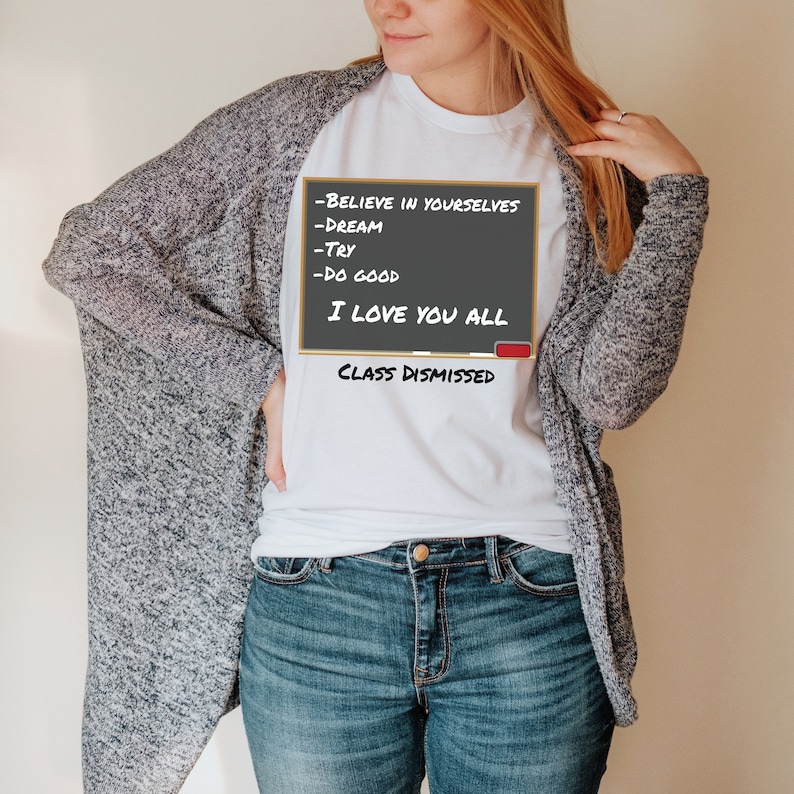 A woman wearing a white t-shirt that has a graphic of a blackboard on it. On the blackboard a lesson is written. It says, Believe in Yourselves, Dream, Try, Do Good. I Love You All. Class Dismissed.
