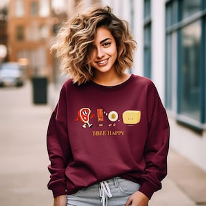 May include: Burgundy sweatshirt with a graphic of cartoon food characters, including a steak, bacon, egg, and butter. The text "BBBE HAPPY" is printed below the graphic.