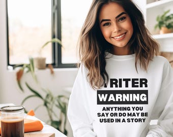 May include: A white sweatshirt with the text "WRITER WARNING" in black, with the phrase "ANYTHING YOU SAY OR DO MAY BE USED IN A STORY" below. The sweatshirt is being worn by a person holding a mug.