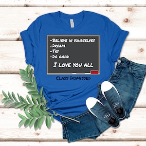 A blue t-shirt that has a graphic of a blackboard on it. On the blackboard a lesson is written. It says, Believe in Yourselves, Dream, Try, Do Good. I Love You All. Class Dismissed.