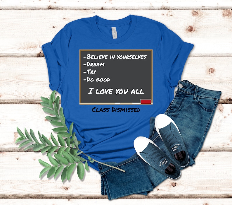 A blue t-shirt that has a graphic of a blackboard on it. On the blackboard a lesson is written. It says, Believe in Yourselves, Dream, Try, Do Good. I Love You All. Class Dismissed.