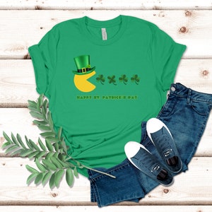 Puede incluir: Camiseta verde con un diseño de Pac-Man que lleva un sombrero de duende verde y come tréboles. El texto "Happy St. Patrick's Day" está impreso debajo del diseño.