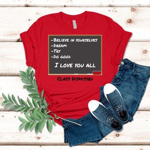 A red t-shirt that has a graphic of a blackboard on it. On the blackboard a lesson is written. It says, Believe in Yourselves, Dream, Try, Do Good. I Love You All. Class Dismissed.