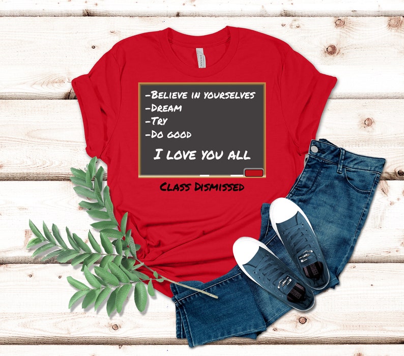 A red t-shirt that has a graphic of a blackboard on it. On the blackboard a lesson is written. It says, Believe in Yourselves, Dream, Try, Do Good. I Love You All. Class Dismissed.