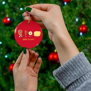 May include: A red, round Christmas ornament with a gold string. The ornament features cartoon illustrations of steak, bacon, an egg, and butter, with the text "BBBE HAPPY" below the illustrations.