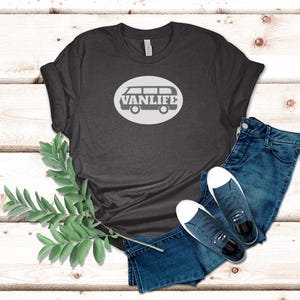 A dark gray t-shirt lays flat. The shirt has a white circular embellishment featuring a graphic of a van that reads VAN LIFE