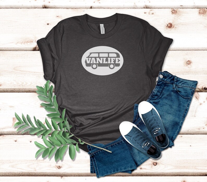 A dark gray t-shirt lays flat. The shirt has a white circular embellishment featuring a graphic of a van that reads VAN LIFE