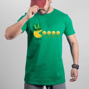 Puede incluir: Camiseta verde con un gráfico de Pac-Man con un sombrero de duende verde y comiendo monedas de oro. El texto "St. Pacman Day" está impreso debajo del gráfico.