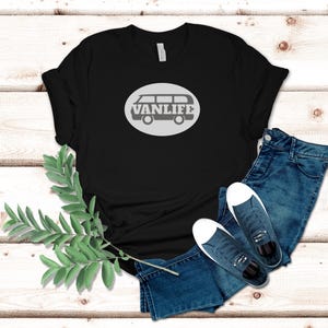 A black t-shirt lays flat. The shirt has a white circular embellishment featuring a graphic of a van that reads VAN LIFE