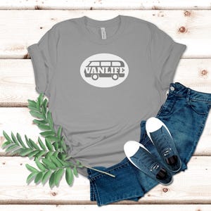 A silver t-shirt lays flat. The shirt has a white circular embellishment featuring a graphic of a van that reads VAN LIFE