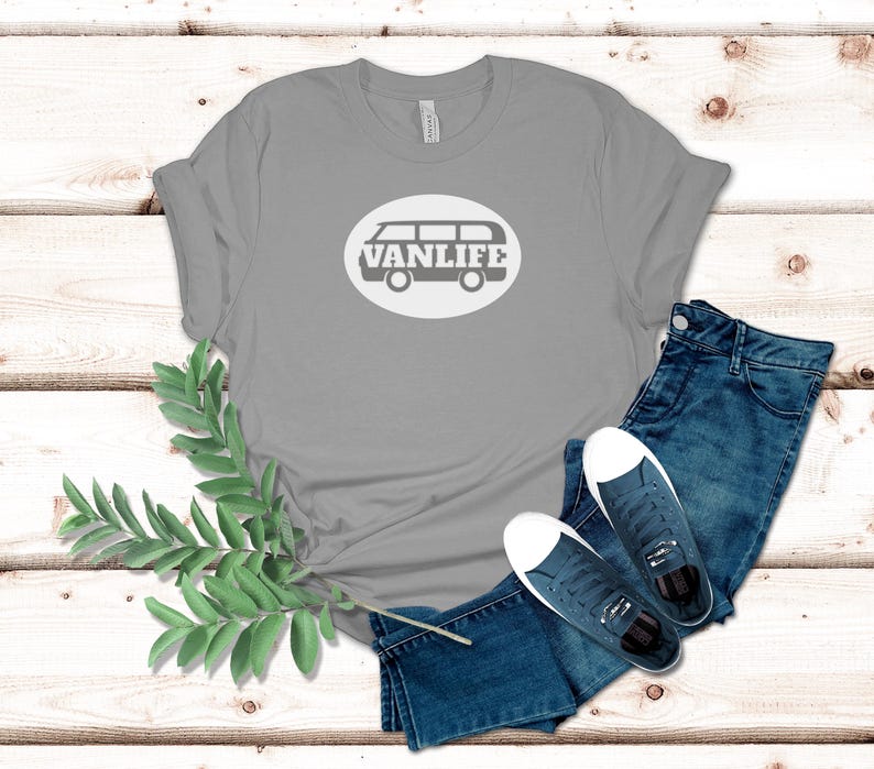A silver t-shirt lays flat. The shirt has a white circular embellishment featuring a graphic of a van that reads VAN LIFE