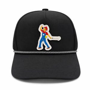 Op de afbeelding: Een zwarte baseballpet met een wit omrande patch van een figuur in een rood shirt en blauwe tuinbroek met de letter N op de borst, die een golfclub vasthoudt. De pet is voorzien van een decoratief zwart-wit gevlochten koord over de rand.