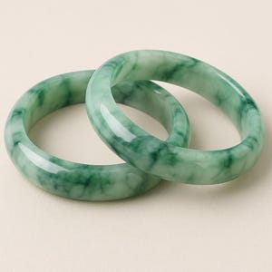 May include: Two green jade bangles with a marbled pattern. The polished bracelets are displayed on a neutral background. These bangles are a classic piece of jewellery.