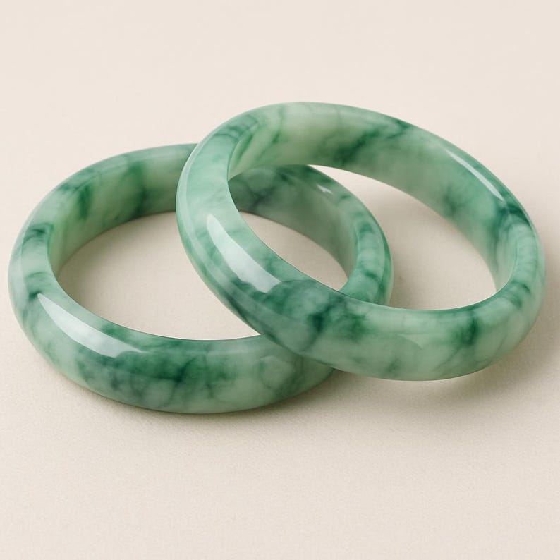 May include: Two green jade bangles with a marbled pattern. The polished bracelets are displayed on a neutral background. These bangles are a classic piece of jewellery.