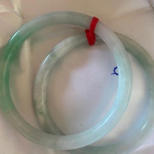 May include: Two jade bangles in shades of green and white are shown. The bangles are round and smooth, with a polished finish. A red string is tied around the bangles. The background is a white, textured surface.