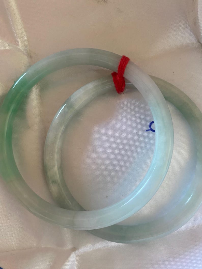 May include: Two jade bangles in shades of green and white are shown. The bangles are round and smooth, with a polished finish. A red string is tied around the bangles. The background is a white, textured surface.