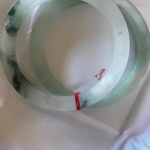 May include: Two jade bangles. The bracelets are a pale green colour with darker green marbling. The bangles are round and smooth, with a polished finish. A red string is wrapped around the bangles.