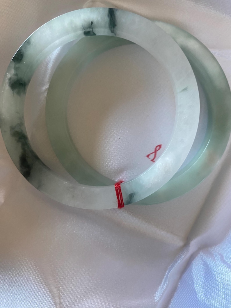 May include: Two jade bangles. The bracelets are a pale green colour with darker green marbling. The bangles are round and smooth, with a polished finish. A red string is wrapped around the bangles.