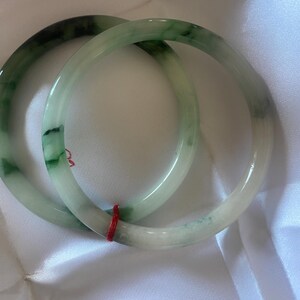May include: Two jade bangles in shades of green and white. The bracelets have a smooth, polished finish and are connected by a red string. The bangles are displayed on a white, silky fabric. Each bangle is approximately 6.5 cm in diameter.