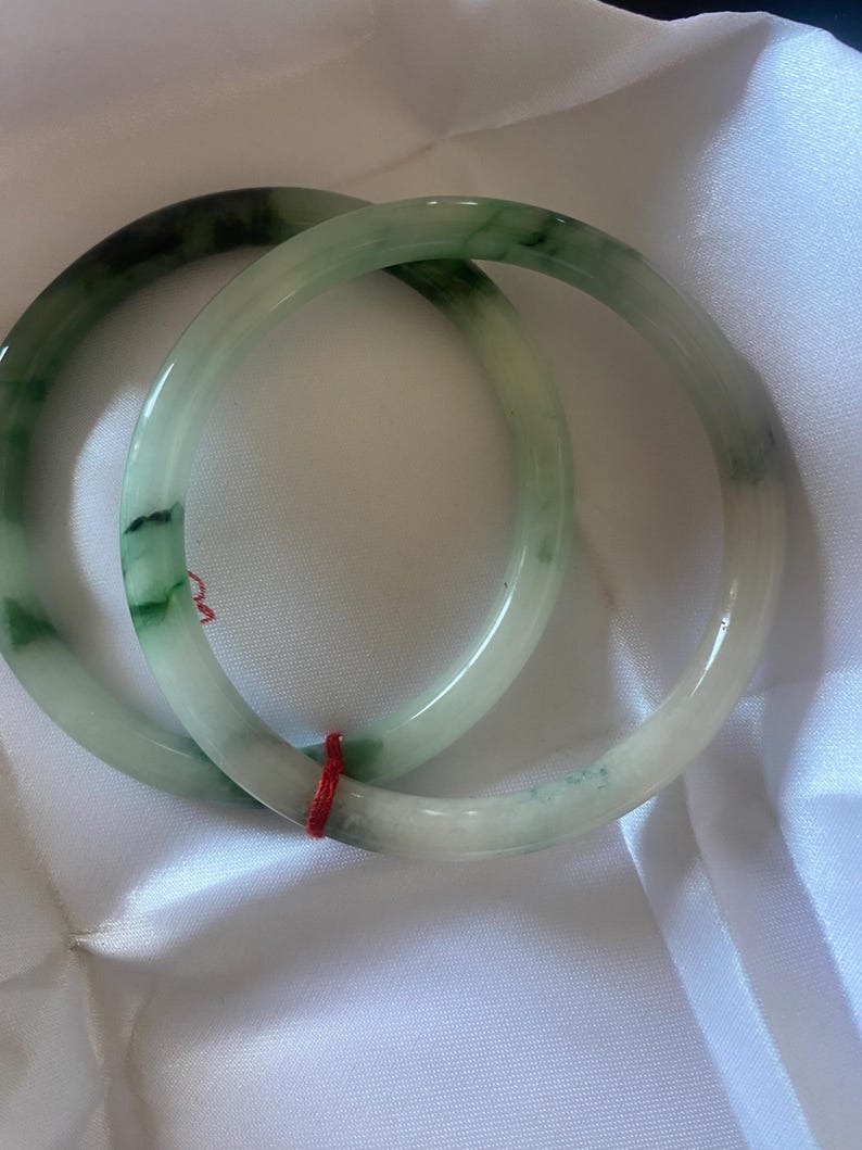 May include: Two jade bangles in shades of green and white. The bracelets have a smooth, polished finish and are connected by a red string. The bangles are displayed on a white, silky fabric. Each bangle is approximately 6.5 cm in diameter.
