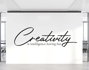 May include: A white wall in an office setting features the black cursive text "Creativity is intelligence having fun." The office has glass-walled rooms and a bright, modern aesthetic. The text is the focal point.