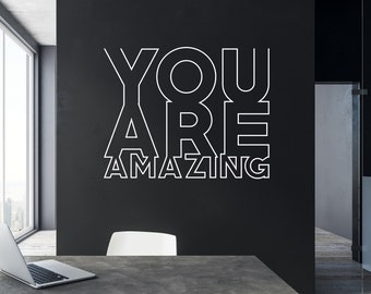 May include: A modern office with a large window overlooking a city skyline. The wall is painted black and features a white vinyl decal that reads "YOU ARE AMAZING". A laptop computer is on a concrete table in the foreground.