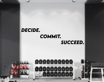 以下が含まれることがあります： 黒い文字で「DECIDE. COMMIT. SUCCEED.」というモチベーションを高める言葉が書かれた白い壁。壁は、黒と白のウェイトラックと黒いウェイトベンチのあるジムにある。