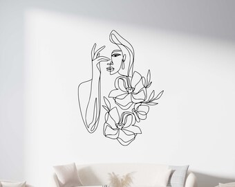 May include: A black and white line drawing of a woman with flowers in her hair. The drawing is on a white wall above a white couch and a coffee table.