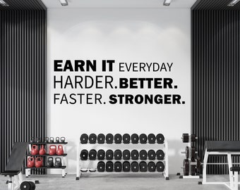 以下が含まれることがあります： 黒い文字で「EARN IT EVERYDAY HARDER. BETTER. FASTER. STRONGER.」というモチベーションを高める言葉が書かれた白い壁。壁は、ウェイトラックとウェイトベンチのあるジムにある。
