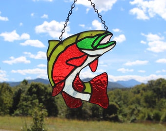 May include: A stained glass fish with red, green, and white accents hanging from a chain. The fish is facing right and has its mouth open. The background is a blurry image of a forest and mountains.