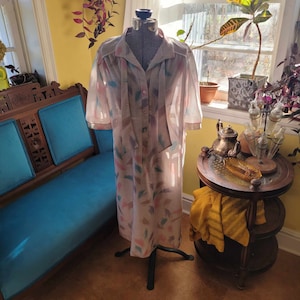 Peut inclure: Une longue robe boutonnée en transparence avec un imprimé floral dans les tons rose, marron et bleu. La robe est sur un mannequin et a un nœud papillon au cou.