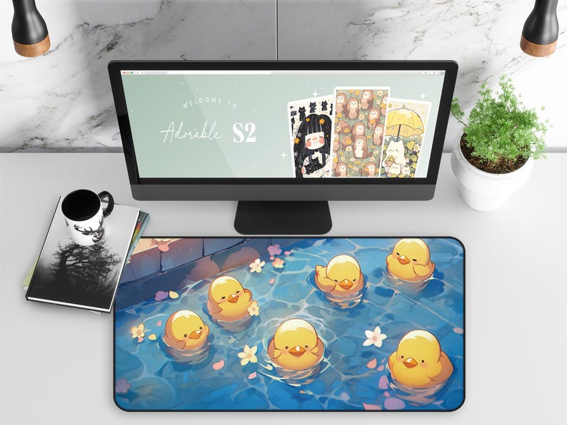 K&ouml;nnte beinhalten: Ein Schreibtisch mit einem Computermonitor, der "Welcome to Adorable S2" und Illustrationen anzeigt. Eine Mausunterlage mit Cartoon-Entenk&uuml;ken im Wasser befindet sich vor dem Monitor.