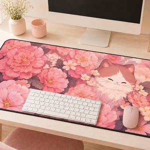 Pode incluir: Um tapete de secretária retangular com um padrão floral de grandes peónias cor-de-rosa e flores brancas mais pequenas. Uma ilustração estilizada de um gato sorridente com pelo castanho e branco está inserida entre as flores à direita.