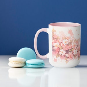 May include: A white ceramic mug with a light pink interior and handle, featuring a printed illustration of pink and white cherry blossoms. Next to the mug are two light blue macarons and one white macaron arranged on a smooth surface.