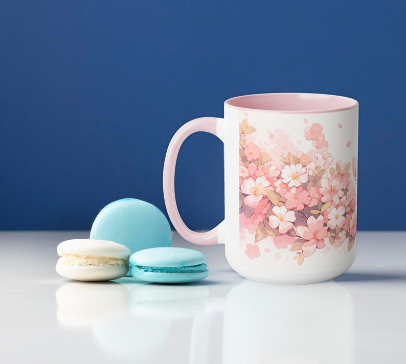 May include: A white ceramic mug with a light pink interior and handle, featuring a printed illustration of pink and white cherry blossoms. Next to the mug are two light blue macarons and one white macaron arranged on a smooth surface.