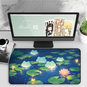 Puede incluir: Una configuración de escritorio con un monitor de computadora que muestra ilustraciones, una alfombrilla de ratón con un diseño de estanque de ranas, una taza, un libro y una pequeña planta en maceta. La alfombrilla de ratón tiene un fondo azul con nenúfares y ranas de dibujos animados.