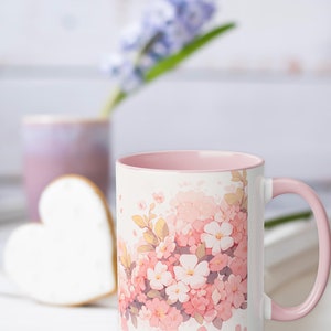 May include: A white ceramic mug featuring a soft pink interior and handle, decorated with a watercolor-style illustration of clusters of pink and white cherry blossoms with small green leaves scattered across the front surface of the mug.