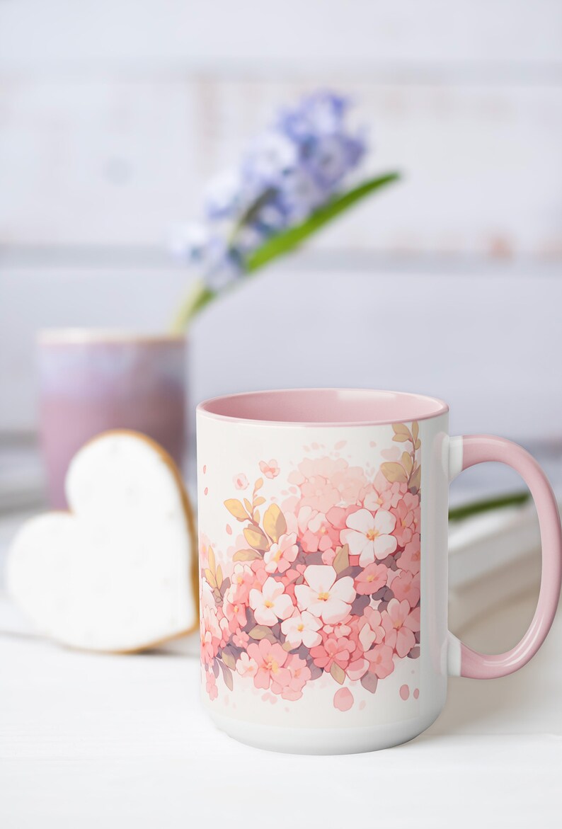 May include: A white ceramic mug featuring a soft pink interior and handle, decorated with a watercolor-style illustration of clusters of pink and white cherry blossoms with small green leaves scattered across the front surface of the mug.