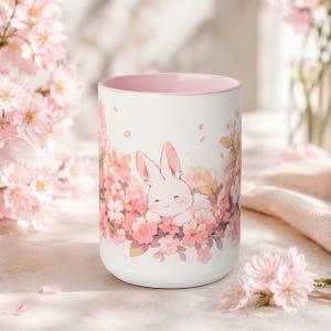 May include: A white ceramic mug with a light pink interior, featuring a graphic illustration of a small white rabbit resting among a cluster of soft pink cherry blossoms. The design wraps around the exterior, showcasing delicate floral petals.
