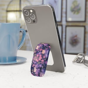 May include: A smartphone propped up by a floral patterned phone grip. The grip features a design of purple and pink flowers with small animal illustrations. A blue mug and framed artwork are in the background.