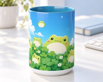 Può includere: Una tazza cilindrica in ceramica con uno sfondo di cielo blu vibrante con nuvole bianche e diverse rane in stile cartoon immerse tra foglie di trifoglio verde e piccoli fiori di margherita bianchi. L'interno della tazza è azzurro.