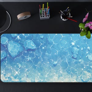 May include: A rectangular desk mat featuring a watercolour design of blue water with white highlights. The mat is surrounded by a black desk, a coffee cup, a lamp, and stationery.