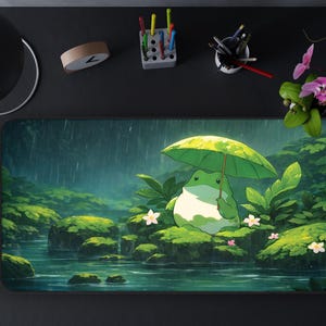 Puede incluir: Alfombrilla de ratón rectangular con una ilustración de una rana bajo un paraguas verde en una escena de bosque lluvioso. La rana es de color verde claro, con un fondo de follaje verde y agua. Flores blancas salpican el paisaje.