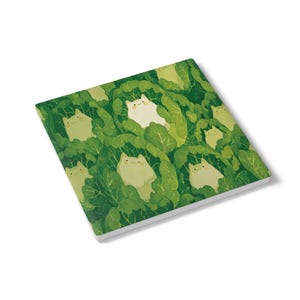 May include: Square notepad featuring a repeating pattern of white cats nestled in green cabbage leaves. The notepad has a white border and is angled slightly, showcasing the design on the top surface.