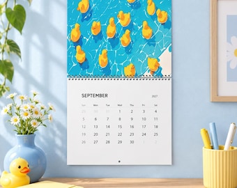 Könnte beinhalten: Ein Wandkalender für September 2027 mit einer Illustration zahlreicher gelber Quietscheentchen, die auf blauem Wasser schwimmen. Der Kalender hat einen weißen Hintergrund mit schwarzem Text für Daten und Wochentage und ist spiralgebunden.