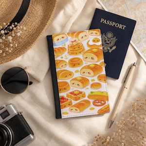 May include: A passport cover with a white background and a pattern of smiling baked goods, including bread, croissants, and pancakes. A dark blue passport and a pen are next to the cover. A straw hat and sunglasses are in the background.