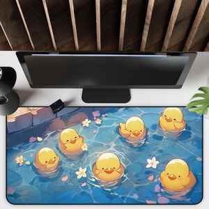 K&ouml;nnte beinhalten: Rechteckige Schreibtischunterlage mit einer Cartoon-Illustration von gelben Entenk&uuml;ken, die in blauem Wasser mit Bl&uuml;tenbl&auml;ttern schwimmen. Die Matte hat einen schwarzen Rand und befindet sich unter einem Computermonitor.